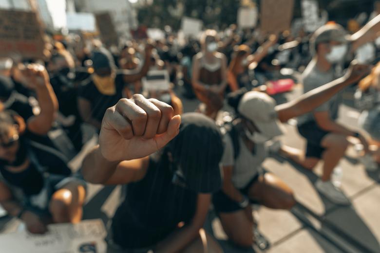 Close up of a black women's fist raised in protest while other protesters take the knee