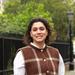 Elifcan has shoulder-length, curly dark hair. She is wearing a white shirt and a brown vest with a large white check. She is standing in front of a park fence, smiling. 