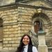Lhori Webster is wearing academic dress and stands in front of a sandstone building in Oxford. 