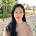 Lin Chen has long dark hair. She is standing in front of some flower beds and foliage and is wearing a beige knitted jumper. 