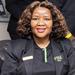 Tlale has curly, shoulder-length brown hair. She is wearing a black polo shirt and blazer, smiling at the camera.