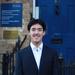 Xin Law-Gallagher stands, smiling, in front of the DSPI blue door. He wears a white shirt and dark blazer. He has short cropped dark hair. 