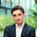 Alessio Cuozzo has short dark hair. He is wearing a white shirt and navy blazer, and is standing in front of a glass building.