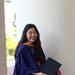 Alexis is sitting on a low wall, smiling. She is wearing a dark blue graduation gown and holding a graduation cap.