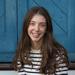 Bridget Steele has long brown hair and is wearing a black and white striped shirt. She is sitting on some outdoor steps in front of a blue door and smiling.