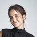 Eliana has brown hair in an updo and is wearing a high-neck draped black top. She is smiling at the camera.
