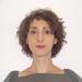 A headshot of Ilka Vari-Lavoisier. She has short curly brown hair. She is standing face-on to the camera, wearing a square-necked grey top with puffed sleeves.