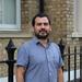 Dr Munoz has short black hair and black facial hair. He is wearing a short-sleeved blue shirt and standing outside a brick building. 