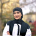Kayanat Iltaf stands with folded arms wearing academic dress in front of an out-of-focus tree in the background. 