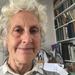 Dr Peretz has short curly white hair. She is sat in front of a large bookcase, smiling. 