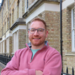Dr Fanner has short fair hair and is wearing glasses. He is standing with his arms crossed, wearing a pink sweater. 