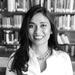 A black and white photo of Rasangi Prematilaka smiling while standing in front of bookshelves. She has long dark hair and is wearing a white shirt and large chain necklace. 