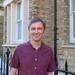 Robin has short fair hair and is wearing a short-sleeved burgundy shirt. He is standing outside in front of some light-brown brick buildings.