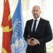 Mr. Benjamin Perks is wearing  a suit and tie standing in front of flags including at UN flag.