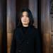 Xinghao li is wearing a black coat, with shoulder length hair looking at the directly at the camera.