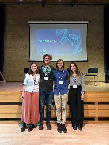 A group of four students standing with their arms around each other, in front of a raised stage and display screen that says 'SERSPI 2024'.