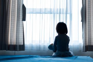 The silhouette of a small child sitting on a bed holding a doll, facing a window with light coming through the curtains.