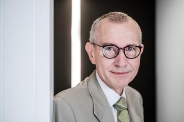 Professor Frank Vandenbroucke is wearing a light suit and round glasses. He is leaning against a light wall.
