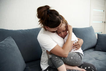 A mother sits on a grey sofa, hugging and comforting her young daughter