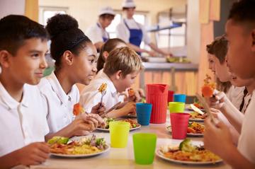 Several primary school children sitting at a lunch table, wearing white polo shirts. They are eating and chatting. The table is filled with multicoloured cups.