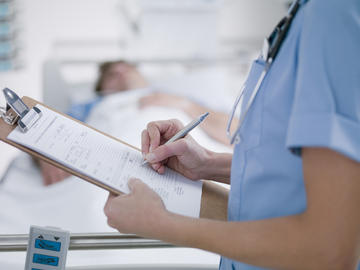 A close up of a nurse writing notes on a clipboard. In the background is a patient in a hospital bed.