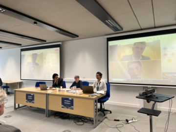 A panel session is taking place in a room, with 3 people sitting in a room with two white screen appearing to be giving a presentation.