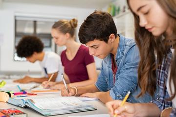 Four young adult students sat in a row at a desk, holding pens and looking at textbooks. 
