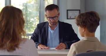 A male teacher sitting at a desk, talking to a mother and child who are sat opposite him with their backs to the camera.