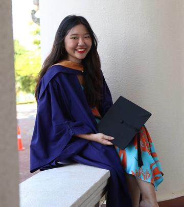 Alexis is sitting on a low wall, smiling. She is wearing a dark blue graduation gown and holding a graduation cap.