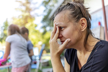 A mother sitting outside, resting her forehead on her fingertips. Children are playing behind her. 