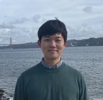 Kun Lee, with short black hair and wearing a green top, smiles at the camera. A bridge and a body of water are in the background.