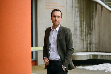 Lukas Lehner wears a white shirt and dark jacket, he stands on an angle next to an orange wall.