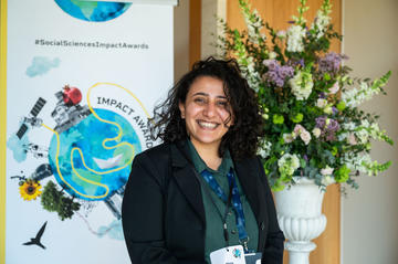 Mona Ibrahim has long curly dark hair. She is smiling broadly, standing in front of a bouquet of flowers and an 'Impact Awards' banner