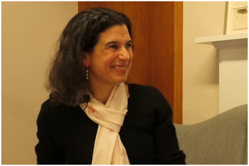 Professor Cluver has long curly black hair. She is wearing a black top and pale pink scarf.