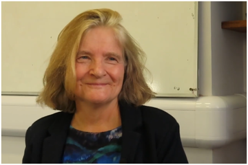 Professor Gardner has short blonde hair. She is wearing a dark blouse with a dark blue and green abstract pattern.