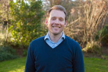 Alex Sutherland has short fair hair and is wearing a light blue shirt with a navy jumper. He is standing outside in a garden, smiling at the camera. 