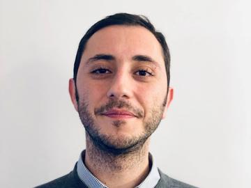 Dr Bedük has short dark hair. He is wearing a light blue shirt and grey jumper. 