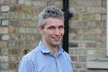 Jonathan smiling at the camera. He has short grey hair and is wearing a blue and white checked shirt. 