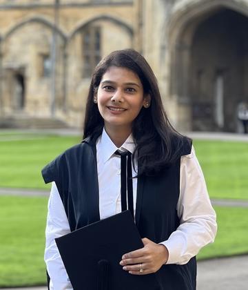 Wajeeha is standing in a large courtyard and smiling at the camera. She has long brown hair and is wearing a black matriculation gown, holding a graduation cap. 