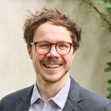 William Rudgard has short brown hair and round red glasses. He is wearing a shirt and blazer and smiling at the camera. 