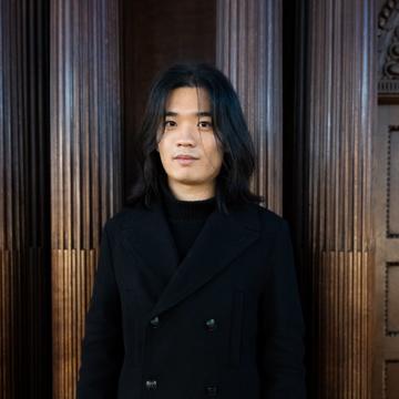Xinghao li is wearing a black coat, with shoulder length hair looking at the directly at the camera.