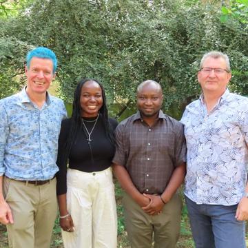 Four people smiling outdoors in front of greenery, standing side by side smiling at the camera.
