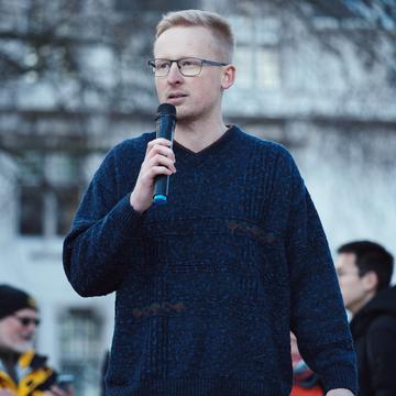 In jeans and a dark blue top, Ben Goodairds is holding a microphone and appears to be speaking outside in a public space.