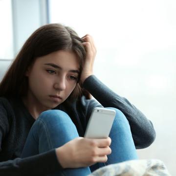 A young girl with long dark hair is sitting on a chair, looking at her phone with a sad expression. 
