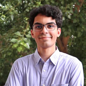 Ansh has curly dark hair and glasses. He is smiling and standing with his arms folded in front of a tree.