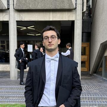 Daniele is wearing glasses a suit, a bow tie, and a graduation gown directly at the camera.
