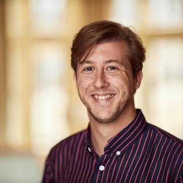 Dirk Witteveen has red hair short hair, wearing a black and red striped shirt smiling at the camera.