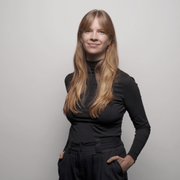 Dr. Bettina Hünteler has long dark-blonde hair, and is standing in front of a plain background wearing a grey polo neck