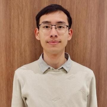 Dr Zhiwu Wei has short dark hair and glasses. He is wearing a beige shirt and jumper and stands in front of a wood-effect wall.