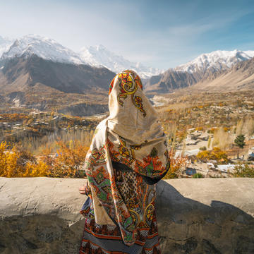 A woman wearing traditional dress sitting on wall and looking at Hunza valley in autumn season, Gilgit Baltistan in Pakistan, Asia